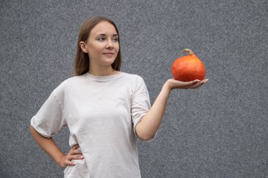Beyaz tişört giyen genç, Avrupalı güzel bir kadın, özel bir tatil için hazırlanıyor, olgun turuncu bir balkabağı tutuyor ve bakışlarını kaçırıyor, gri arka planda izole bir şekilde. Cadılar Bayramı partisi ve insanların konsepti.