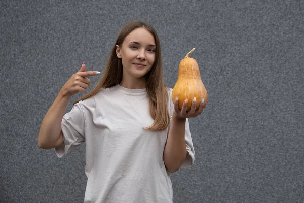 Beyaz tişörtlü genç bir kız, elinde balkabağı, parmağıyla onu işaret ediyor ve gri arka plandaki kameraya bakıyor. Cadılar Bayramı Dekorasyonu, Sonbahar, Kutlama.