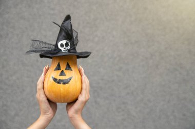 Yatay çekim, şapkada turuncu balkabağı tutan kadın eli ya da gri arka planda balkabağı. Şükran Günü Cadılar Bayramı Boş kopyalama alanı.