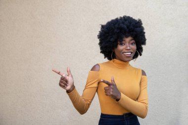 Kıvırcık saçlı mutlu Afrikalı kadın, kameraya bakıyor, fotokopi alanını gösteriyor, hoş bir gülümsemesi var, günlük kıyafetler giyiyor, boş alanda reklam yapıyor, açık arka planda poz veriyor..