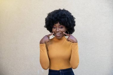Kıvırcık saçlı ve neşeli yüzlü mutlu Afrikalı bir kadının yatay görüntüsü, geniş gülümsemeye işaret ediyor, günlük giyiniyor, beyaz mükemmel dişlerini gösteriyor ve kamera karşısında açık arkaplanda poz veriyor..