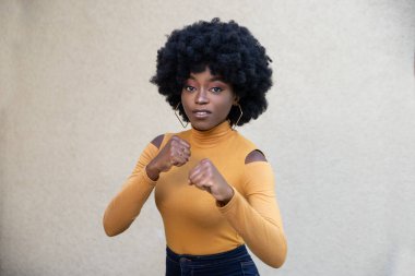 Kıvırcık saçlı güzel bir Afro-Amerikalı kadın, sıkılmış yumruklarını kaldırıyor, mücadele etmeye hazır, gündelik giysiler giyiyor, açık arka planda poz veriyor. Enerjik kız hareketleri..