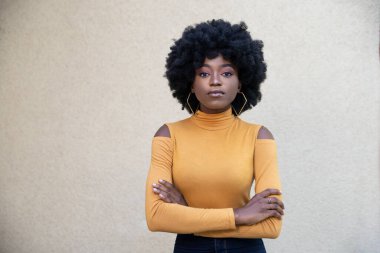 Konsantre olmuş kıvırcık saçlı, kendine güveni tam genç bir Afro-Amerikalı kadın ellerini göğsünün üstüne koyuyor, günlük giysiler giyiyor, kameraya bakıyor, arka planda poz veriyor. Halk konsepti.