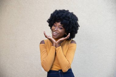 Kıvırcık saçlı ve mutlu yüzlü Afro-Amerikan bir kadın, avuçlarını tutuyor, çenesini duyuyor, rahat giyiniyor, kameraya bakıyor, açık arkaplanda boş fotokopi alanı ile poz veriyor..