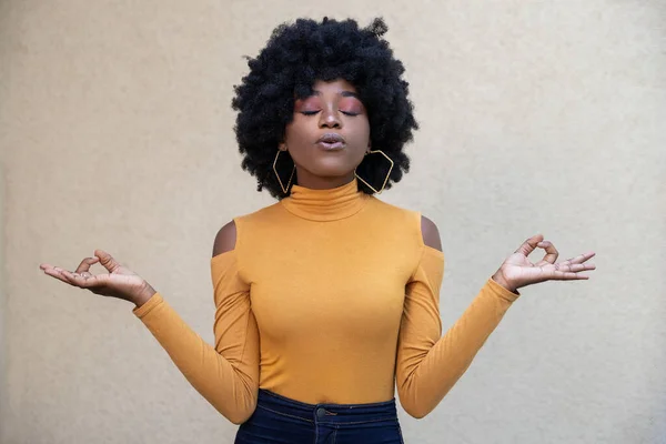 Horizontal shot of calm relieved African American woman with curly hair and closed eyes, keeps hands sideways in zen gesture, practices yoga and relaxation, posing outdoors on light background.