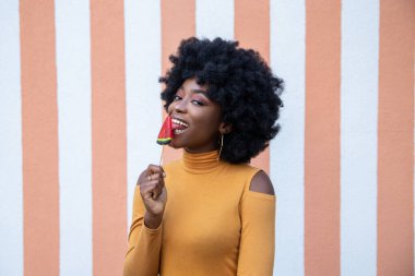 Pozitif esmer, kıvırcık saçlı, lolipopu yalayan, tatlı seven, sıradan kıyafetler giyen, çizgili arka planda poz veren genç bir kadının portresi..