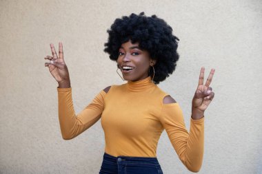 Kıvırcık saçlı, pozitif Afro-Amerikan kadın, huzur ya da zafer işareti gösteriyor, günlük giysiler giyiyor, geniş bir gülümseme ve açık arka planda açık havada poz veriyor..