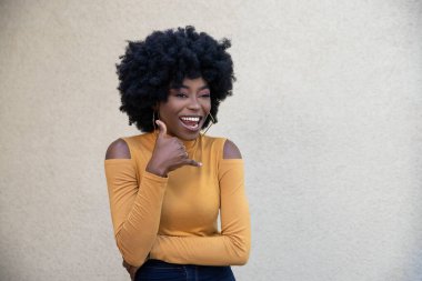 Kıvırcık saçlı, neşeli yüzlü, arkadaş canlısı Afrikalı Amerikalı bir kadının yatay çekimi, telefon işareti gösteriyor, arkadaşlarıyla görüşüyor, günlük giysiler giyiyor, açık arka planda poz veriyor..