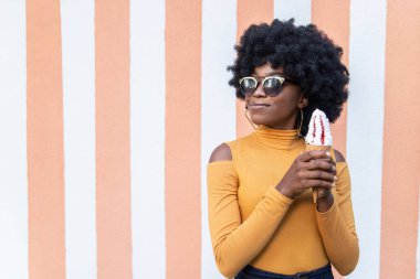 Kıvırcık saçlı, güneş gözlüklü, çilekli dondurma külahında dondurma tutan, sarı balıkçı yaka giyen, çizgili arka planda poz veren çekici Afrikalı kadın. Boş kopyalama alanı.