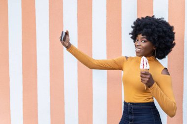 Kıvırcık saçlı, mutlu, esmer bir kadın, kameralı telefondan selfie çekiyor, waffle külahında lezzetli bir dondurma tutuyor, çizgili arka planda poz veriyor. Boş kopyalama alanı. 