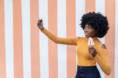 Kıvırcık saçlı komik Afrikalı Amerikalı kadın, akıllı telefonuyla kendi portresini çekiyor, çilekli dondurma külahında, çizgili arka planda poz veriyor. Boş kopyalama alanı.