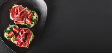 Bütün tahıl ekmeği sandviç jambon, avokado, salatalık, domates, otlar ve tohumlar siyah taş zemin üzerine plaka ile. Temiz yemek, sağlıklı kahvaltı. Üstten görünüm, düz yatıyordu