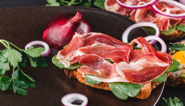 Bütün tahıl ekmeği sandviç jambon, avokado, salatalık, domates, otlar ve tohumlar siyah taş zemin üzerine plaka ile. Temiz yemek, sağlıklı kahvaltı. Üstten görünüm, düz yatıyordu