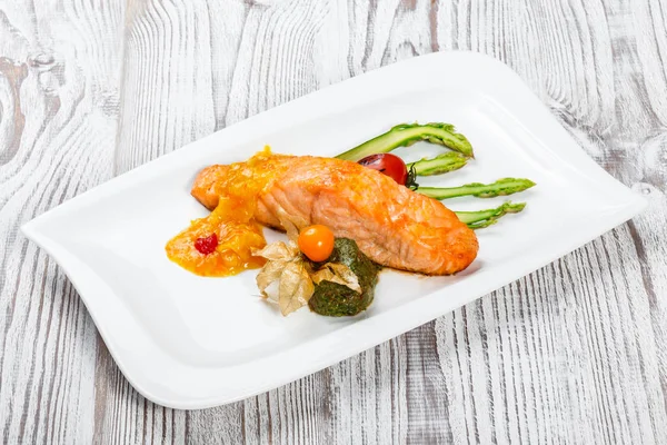Baked salmon garnished with physalis, asparagus, tomatoes with herbs on wooden background. Hot fish dish. Top view