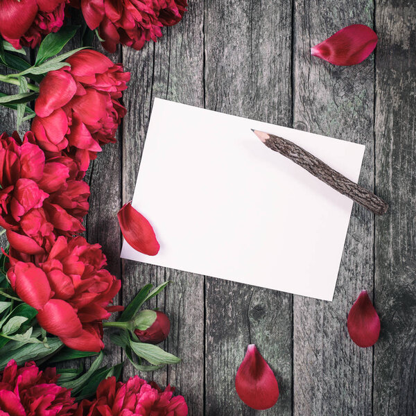 Pink peonies flowers and sheet of paper over dark wooden background with space for text. Mother's Day frame and spring background concept. Greeting card or wedding invitation. Flat lay, top view