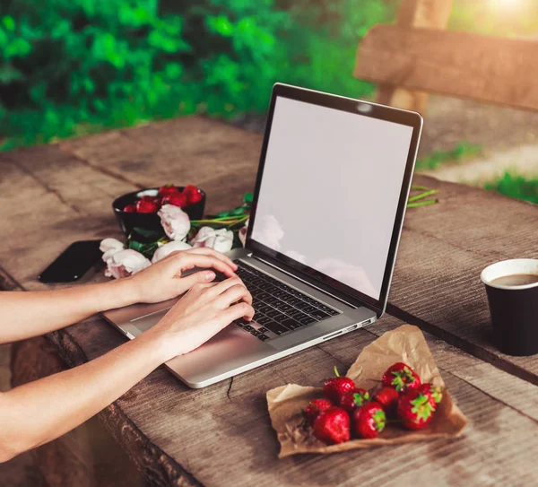 stock image Young woman using and typing laptop computer at rough wooden table with coffee cup, strawberries, bouquet of peonies flowers.