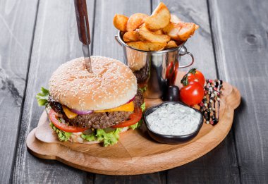Hamburger taze sebze, peynir, sos ve patates kızartması kesme tahtası üzerinde koyu ahşap arka plan üzerinde. Burger bıçakla. Amerikan fast food
