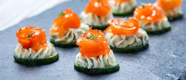Appetizer canape with salmon, cucumber and cream cheese on stone slate background close up. Delicious snacks, sandwiches, crostini, bruschetta, antipasti on party or picnic time. Top view