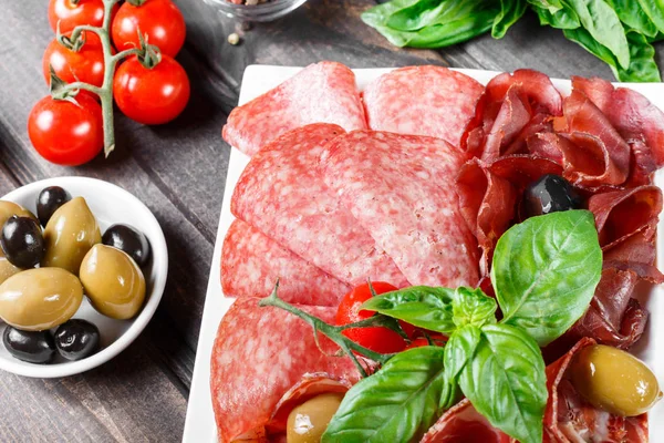 Antipasto platter cold meat plate with prosciutto, slices ham, salami, decorated with basil and olive on wooden background. Meat appetizer. Top view