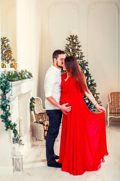 Happy family couple on Christmas at fireplace. Happy couple kiss and hugging. Living room decorated by Xmas tree and present gift box, the light give cozy atmosphere. Waiting for baby. New Year theme. Toning