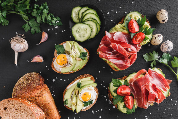 Разнообразные сэндвичи и брускетта с прошутто, жареным перепелиным яйцом, авокадо, огурцом, помидорами, специями и зеленью на черном каменном фоне. Чистое питание, здоровый завтрак. Вид сверху, плоский
