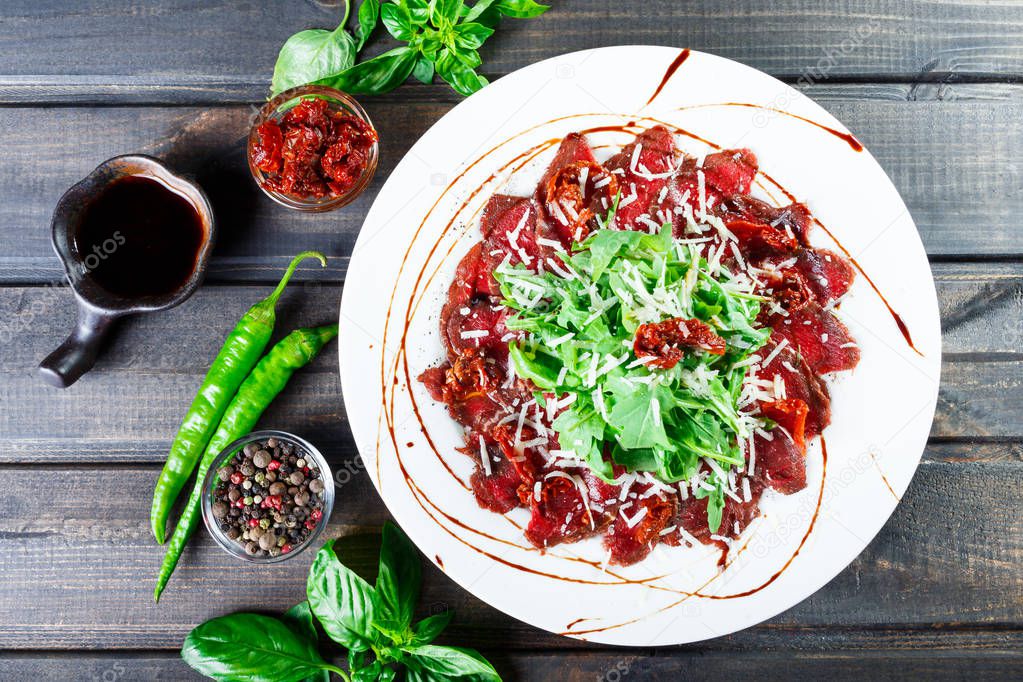 Beef carpaccio with arugula, sun-dried tomatoes, and cheese parmesan on dark wooden background. Delicious appetizers. Top view.