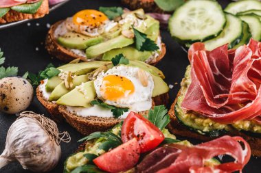 Çeşitli sandviçler ve jambon, kızarmış bıldırcın yumurta, avokado, salatalık, domates, baharatlar ve siyah taş zemin üzerine yeşil ile bruschetta. Temiz yemek, sağlıklı kahvaltı. Üstten görünüm, düz yatıyordu