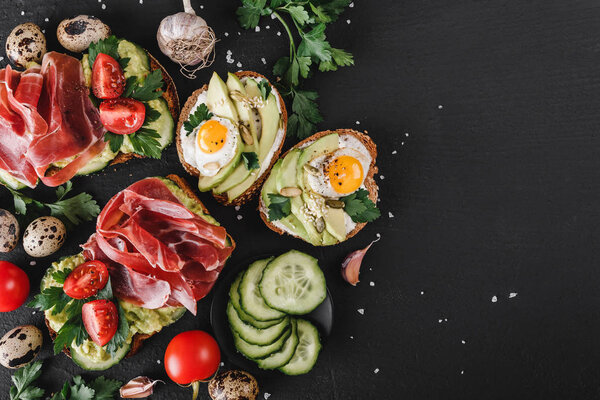 Разнообразные сэндвичи и брускетта с прошутто, жареным перепелиным яйцом, авокадо, огурцом, помидорами, специями и зеленью на черном каменном фоне. Чистое питание, здоровый завтрак. Вид сверху, плоский
