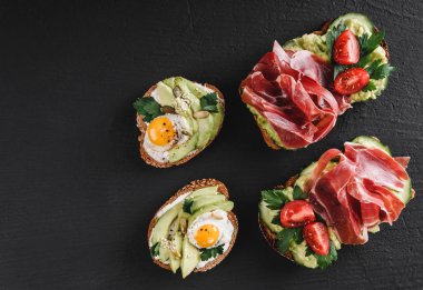Çeşitli sandviçler ve jambon, kızarmış bıldırcın yumurta, avokado, salatalık, domates, baharatlar ve siyah taş zemin üzerine yeşil ile bruschetta. Temiz yemek, sağlıklı kahvaltı. Üstten görünüm, düz yatıyordu