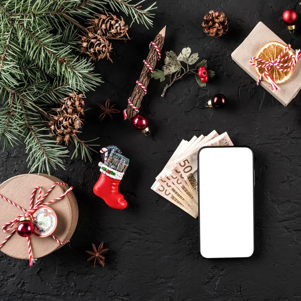 Christmas composition with mobile phone, money euro, Fir branches, gifts, red decorations on dark background. Xmas and Happy New Year theme. Flat lay, top view