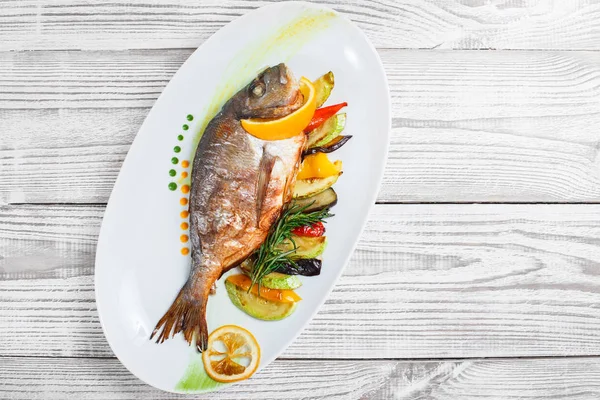 Grilled dorado fish with baked vegetables and rosemary on plate on wooden background close up. Healthy food. Top view