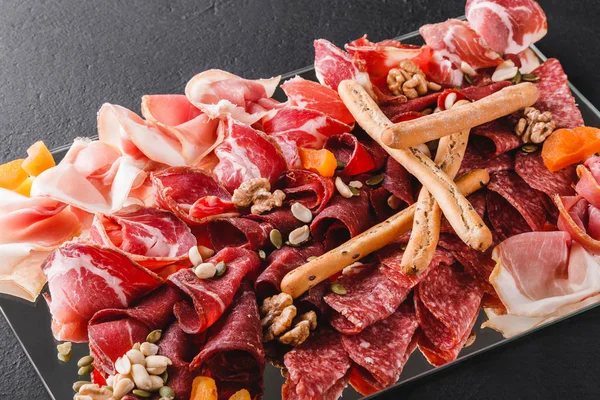 Antipasto platter cold meat with grissini bread sticks, prosciutto, slices ham, beef jerky, salami, meat and nuts on black stone background. Meat appetizer, catering food concept