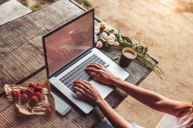 Kahve fincanı, çilek, şakayık çiçeği ve akıllı telefonlu kaba ahşap masada dizüstü bilgisayar kullanan ve daktilo kullanan genç bir kadın. Açık hava parkında çalışan serbest çalışan biri. Tonlama
