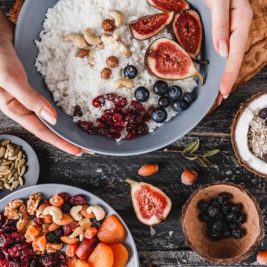 Tabak pilav Hindistan cevizi lapası incir, çilek, fındık ve Hindistan cevizi sütü ile rustik ahşap arka plan üzerinde tutan kadın. Sağlıklı kahvaltı malzemeler. Yeme, vegan gıda kavramı temiz. Üstten görünüm