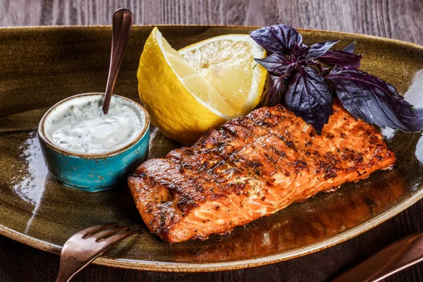 Baked salmon fillet with cheese sauce, basil and lemon on plate on wooden background. Hot fish dish. Top view