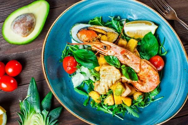 Warm salad with seafood, langoustine, mussels, shrimps, squid, scallops, mango, pineapple, avocado, arugula and basil on wooden background close up. Mediterranean food. Top view