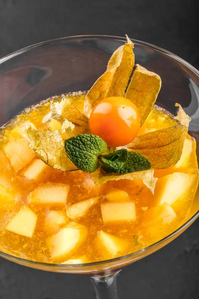 Fruit jelly from mango and peach decorated with mint and physalis in a glass on dark stone background. Summer dessert