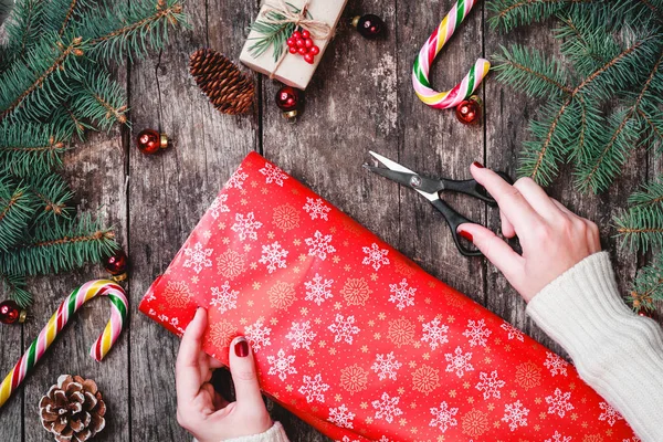 Female hand wraps Christmas gift on holiday wrapping on wooden background with Fir branches, pine cones, red decorations. Xmas and Happy New Year composition. Flat lay, top view