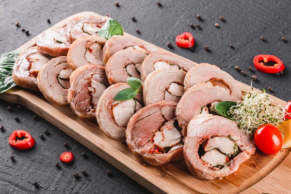 Assorted meat, stuffed chicken roll, meat roll with pepper, greens on cutting board on black shale background. Meat appetizer, food concept