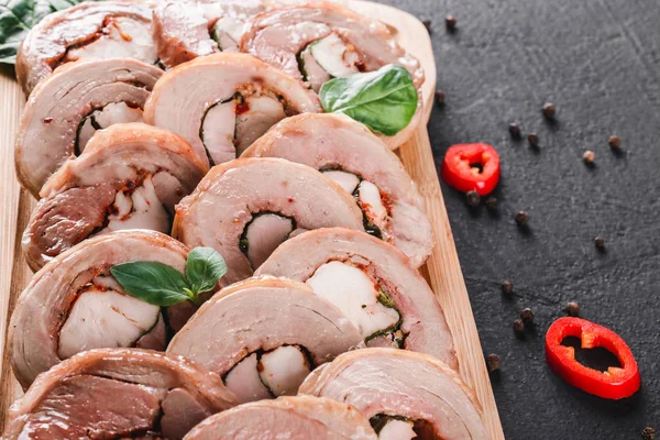 Assorted meat, stuffed chicken roll, meat roll with pepper, greens on cutting board on black shale background. Meat appetizer, food concept