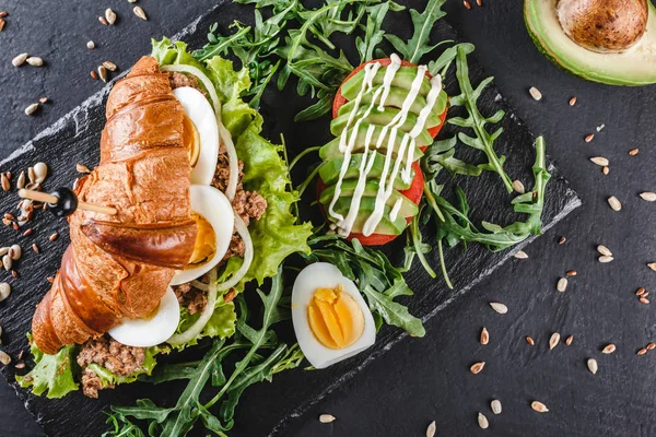 Sandwich with tuna, eggs, avocado, fresh arugula and greens on black shale board over black stone background. Healthy food concept. Top view, flat lay