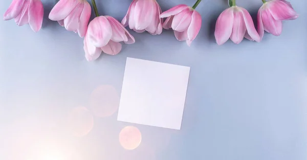 Pink tulips flowers and sheet of paper over light blue background. Card for Mothers day, 8 March, Happy Easter. Greeting card or wedding invitation. Flat lay, top view
