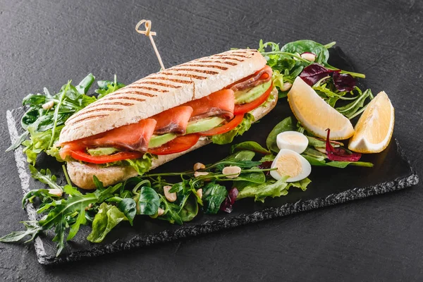 Sandwich con pescado rojo, huevos, aguacate, verduras frescas y verduras en pizarra de esquisto negro sobre fondo de piedra negra. Concepto de comida saludable — Foto de Stock