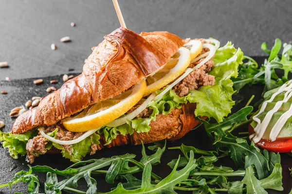 Sandwich de croissant con atún, aguacate, rúcula fresca y verduras sobre pizarra negra sobre fondo de piedra negra. Concepto de comida saludable — Foto de Stock