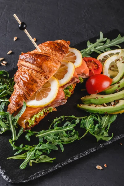 Sandwich mit rotem Fisch, Avocado, frischem Gemüse und Rucola auf schwarzem Schieferbrett vor schwarzem Steinhintergrund. gesundes Ernährungskonzept — Stockfoto