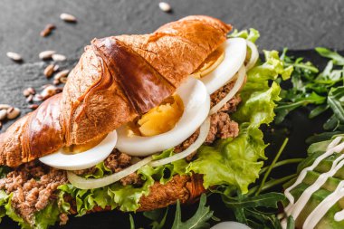 Siyah taş arka plan üzerinde siyah Shale tahta üzerinde ton balığı, yumurta, avokado, taze roka ve yeşillikler ile kruvasan sandviç. Sağlıklı gıda konsepti