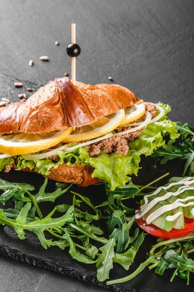 Croissant-Sandwich mit Thunfisch, Avocado, frischem Rucola und Grüntönen auf schwarzem Schieferbrett vor schwarzem Steinhintergrund. gesundes Ernährungskonzept — Stockfoto