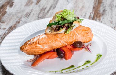 Ahşap arka plan üzerinde tabak üzerinde zeytin, yeşillik, domates ile süslenmiş pişmiş somon. Sıcak balık yemeği.