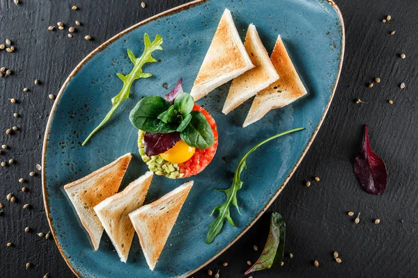 Sığır tartar bir yumurta sarısı, doğranmış avokado, domates ve tost koyu taş arka planda plaka ile hizmet vermiştir. Temiz yemek, sağlıklı gıda kavramı. Üst görünüm