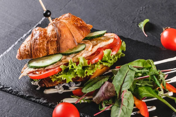 Sandwich de croissant con filete de pollo a la parrilla, verduras frescas, queso y verduras en pizarra negra sobre fondo de piedra negra. Concepto de comida saludable — Foto de Stock